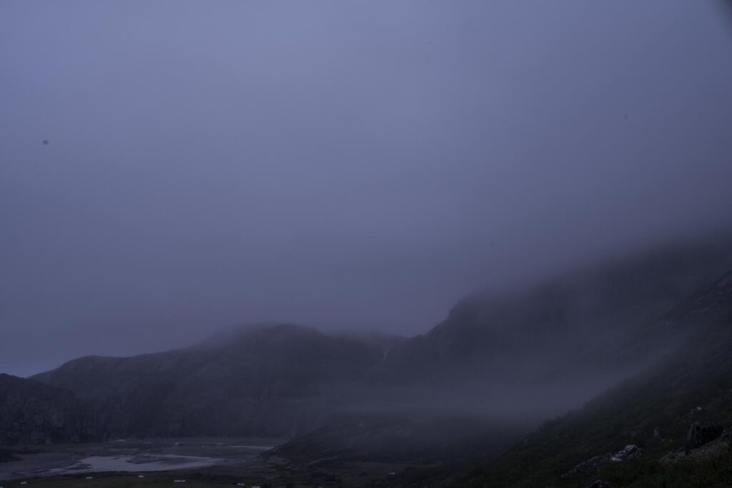 A dense fog covering a valley floor between mountain ranges, with only the faintest outlines of the arctic Greenland terrain visible