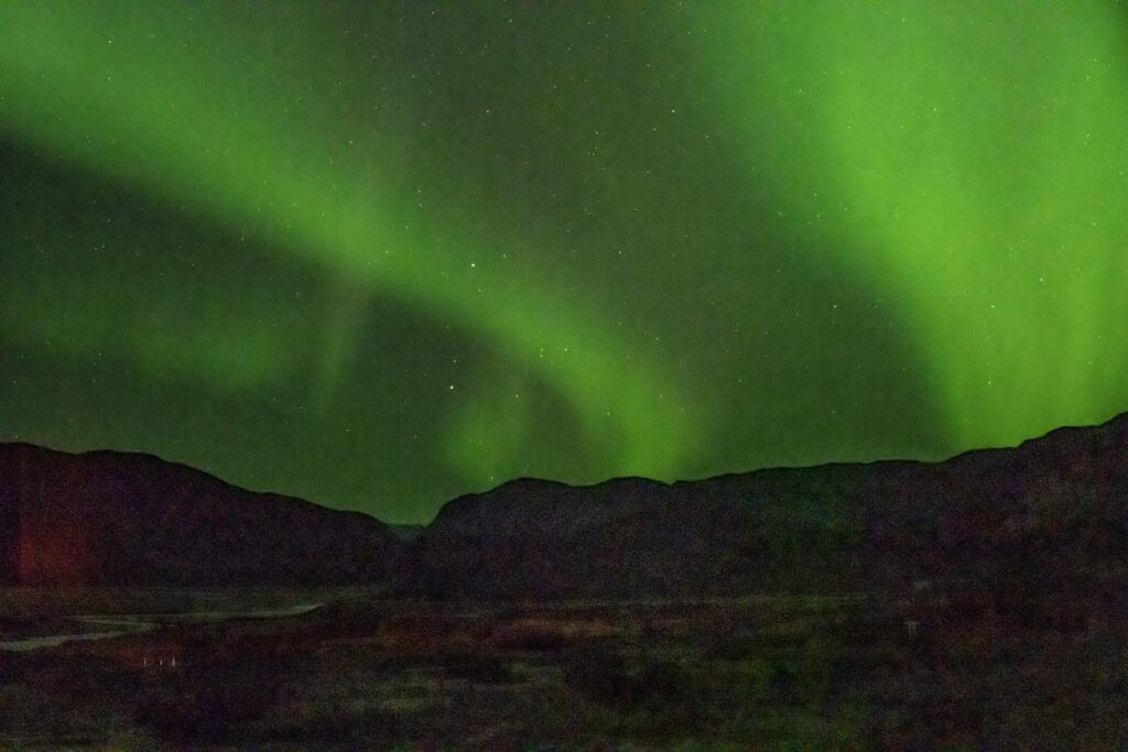 Star trails arcing through the night sky above a dark landscape with silhouetted mountains and a lit-up valley floor in Greenland