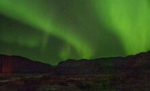 Star trails arcing through the night sky above a dark landscape with silhouetted mountains and a lit-up valley floor in Greenland