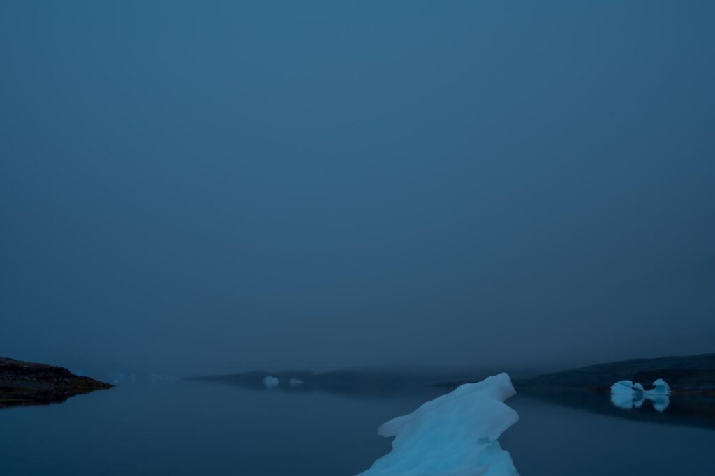 A luminous, sculptural iceberg floating in dark, still water against a moody, overcast sky in Southern Greenland, photographed by Steve Giovinco.